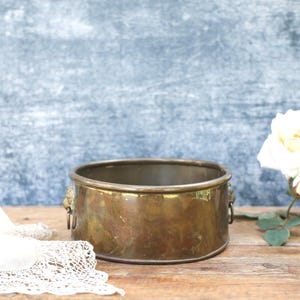 May include: A round, polished brass bowl featuring two decorative lion head handles on opposite sides. The metal surface shows natural patina and variations in colour, with a smooth, wide rim and a flat base, presenting a classic, functional design.