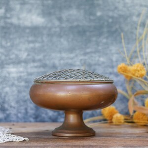 May include: A vintage copper flower frog with a metal grid lid. The container has a rounded body and a pedestal base. Dried yellow flowers and a lace doily are in the background.