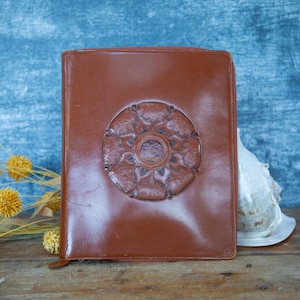 May include: A brown leather-bound book with a decorative embossed floral design on the cover. The book is rectangular and appears to be vintage, with a worn appearance. The background is a textured blue surface.