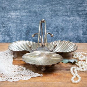 May include: A silver-toned, three-sectioned serving dish with a handle, shaped like seashells. A white lace doily and a strand of faux pearls are on a wooden surface. The background is a textured blue.