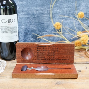 May include: A dark glass wine bottle labelled CARO next to an open rectangular wooden box containing a metal wine thermometer. The inside lid of the box features engraved text listing various wine types and their recommended serving temperatures.