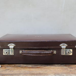 May include: A rectangular dark brown hard-shell suitcase featuring a central leather handle, two silver-toned metal latches with integrated keyholes, and reinforced silver-toned metal corners. The exterior surface shows signs of wear.