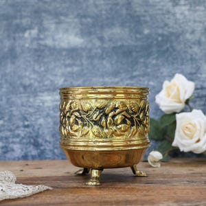 May include: A decorative gold-coloured metal planter with a floral pattern. The planter is supported by three small feet and is displayed on a wooden surface. White roses are in the background.