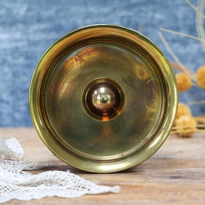 May include: A circular brass candle holder featuring a wide, flat rim and a central, raised dome-shaped spike designed to secure a candle. The polished metal surface exhibits a warm, golden metallic tone with subtle variations in its finish.