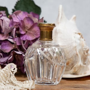 May include: A clear, faceted glass perfume bottle with a gold-colored cap. The bottle is round and sits on a wooden surface, next to a large seashell and purple hydrangea flowers. A vintage aesthetic.