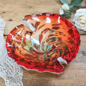 May include: A decorative glass bowl featuring a vibrant swirl of red, orange, and dark brown colours with a glossy finish. The bowl has an irregular, organic shape with a ruffled edge and a smooth, reflective surface that catches the light.