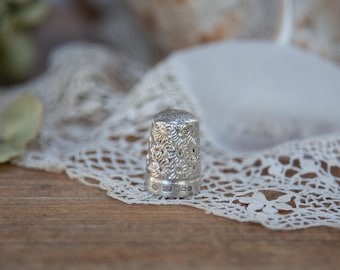 May include: A silver thimble with an intricate floral pattern. The thimble is cylindrical with a slightly rounded top and small holes near the top. The base of the thimble has markings. The thimble is set on a white lace doily.