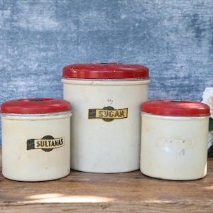 May include: Three vintage cream-coloured canisters with red lids. The largest canister is labelled "Sugar", while the smaller ones are labelled "Sultanas" and "Spice". The canisters are arranged on a wooden surface.