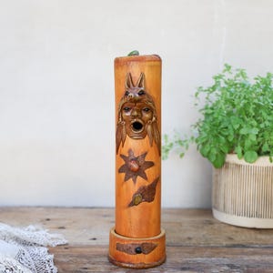 May include: A cylindrical wooden sculpture featuring a carved face wearing an animal headdress, a star shape, and a small leaf motif. The light brown wood has a smooth finish and sits on a circular base, showcasing detailed relief carvings.