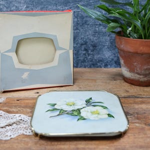 May include: A square trivet with a light blue background and a white floral illustration featuring two blossoms with green leaves. The trivet has a thin gold-toned metal frame and is accompanied by a cardboard box with a geometric cutout design.
