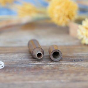 May include: Two small, cylindrical wooden objects with a dark brown finish. One has a hole at the end, and the other has a hollowed-out interior. The items are on a wooden surface, with yellow floral accents in the background.