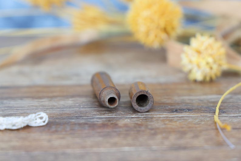 May include: Two small, cylindrical wooden objects with a dark brown finish. One has a hole at the end, and the other has a hollowed-out interior. The items are on a wooden surface, with yellow floral accents in the background.