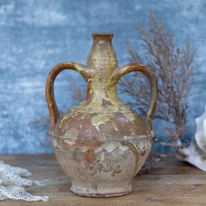 May include: A rustic, two-handled ceramic vase with a weathered appearance. The vase has a bulbous body and a narrow neck, with two arched handles. The color is a mix of brown, tan, and yellow, with visible signs of wear. The vase is likely an antique or vintage piece.