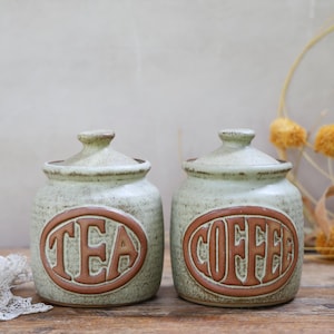 May include: Two ceramic tea and coffee storage jars with lids. The jars are a speckled green colour with brown lettering and trim. The jar labelled "TEA" is next to the jar labelled "COFFEE".