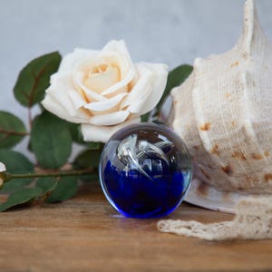 Pode incluir: Um peso de papel de vidro azul com um golfinho prateado dentro, sentado em uma superfície de madeira com uma concha e rosas brancas.