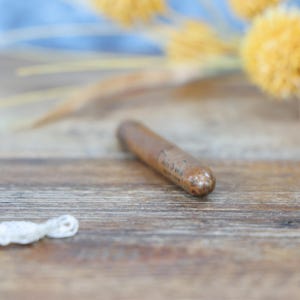 May include: A cylindrical, brown wooden object with a rounded tip, possibly a bottle stopper, rests on a wooden surface. The object has a slightly textured appearance. Dried yellow flowers and a white lace trim are in the background.
