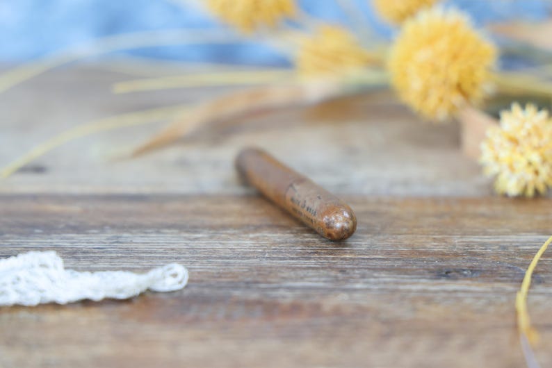 May include: A cylindrical, brown wooden object with a rounded tip, possibly a bottle stopper, rests on a wooden surface. The object has a slightly textured appearance. Dried yellow flowers and a white lace trim are in the background.