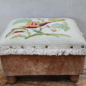 May include: A square footstool with a light beige needlepoint top featuring a floral pattern in green, yellow, and red. The base is made of light brown wood with four short legs. A decorative trim with fringe and metal studs edges the top.