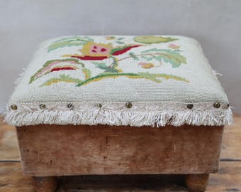 May include: A square footstool with a light beige needlepoint top featuring a floral pattern in green, yellow, and red. The base is made of light brown wood with four short legs. A decorative trim with fringe and metal studs edges the top.