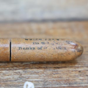 May include: A vintage wooden pencil case with the text "WHICH GREW On the Banks of - - -" printed on the side. The case is a light brown color and has a cylindrical shape with a split in the middle.