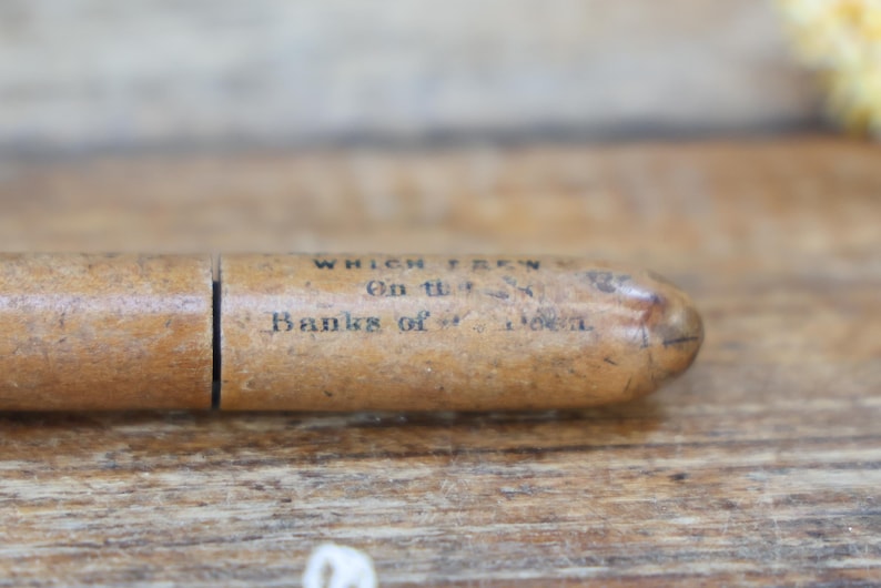 May include: A vintage wooden pencil case with the text "WHICH GREW On the Banks of - - -" printed on the side. The case is a light brown color and has a cylindrical shape with a split in the middle.