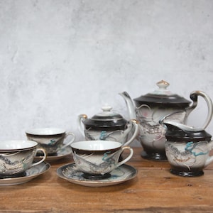 Op de afbeelding: Een vintage porseleinen theeservies met een drakenmotief. De set bevat een theepot, suikerpot, melkkan en twee theekopjes met schotels. De stukken zijn voornamelijk wit met zwarte accenten en drakenillustraties.