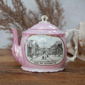 May include: A pink ceramic teapot with a lid, spout, and handle. The teapot features a black and white illustration of a street scene with the text "ABOVE BAR SOUTHAMPTON". Gold trim accents the edges.