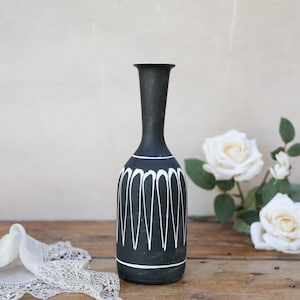 May include: A tall, slender black vase with a matte finish, featuring a white hand-painted pattern of vertical, elongated petal-like shapes around the body and a single horizontal white line near the base, with a narrow neck and flared rim.