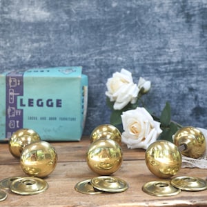 May include: A set of polished brass spherical door knobs with matching circular plates and a light blue Legge brand box. The box features the text LEGGE LOCKS AND DOOR FURNITURE. Two white artificial roses and a piece of white lace are also present.