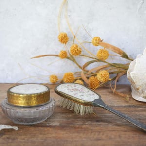 May include: An antique vanity set featuring a small, round jar with a gold rim and a decorative brush with a floral design. The set is placed on a wooden surface with dried yellow flowers and a seashell in the background.