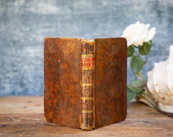 May include: An old book with a distressed, brown leather cover. The spine features gold detailing and red lettering. The book is open, showing aged pages. A white rose and seashell are in the background.