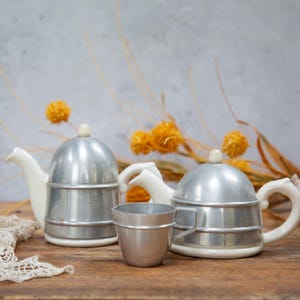 May include: Two vintage espresso makers with white handles and a small metal cup. The espresso makers have a silver metal top and base. The background includes dried orange flowers and a seashell.