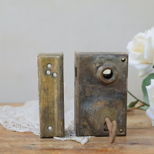 May include: A weathered brass rim lock set featuring a rectangular strike plate with three screw holes and a larger lock body with a circular knob opening and a keyhole containing a small metal key, showing a dark, aged patina throughout.