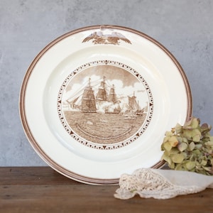 Puede incluir: Un plato de cerámica blanco con un borde marrón y una imagen marrón de tres barcos de vela en el centro. La imagen está rodeada por un diseño de brújula marrón.