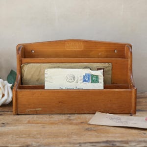 May include: A light brown wooden wall-mounted mail organiser with two tiers. The organiser holds two envelopes, one with a visible postmark and green stamps. The word NATURAL is embossed on the top centre. Two white artificial roses sit to the left.