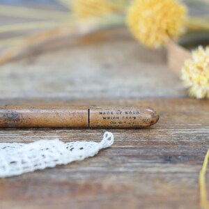 May include: A vintage wooden pencil holder with the text "MADE OF WOOD WHICH FELL" printed on it. The pencil holder is brown and oval-shaped. A piece of white lace is in the foreground, with yellow flowers and stems in the background.