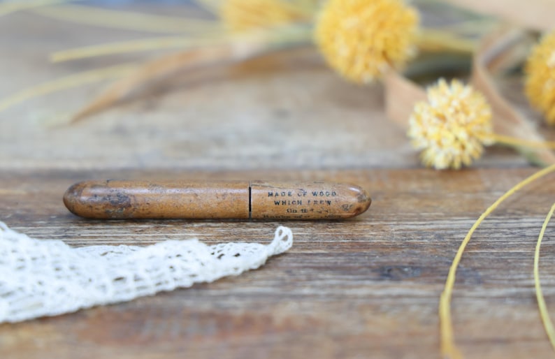 May include: A vintage wooden pencil holder with the text "MADE OF WOOD WHICH FELL" printed on it. The pencil holder is brown and oval-shaped. A piece of white lace is in the foreground, with yellow flowers and stems in the background.