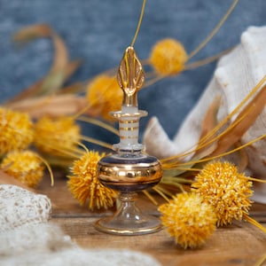 May include: A vintage perfume bottle with a gold and blue design, topped with a gold leaf-shaped stopper. The bottle is surrounded by yellow pom-pom flowers, dried grasses, and a seashell, creating a decorative display.