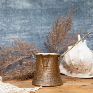 May include: A copper-coloured Turkish coffee pot with a long handle, set on a wooden surface. A large white seashell and dried plants are in the background. The pot has a hammered texture and a decorative rim.
