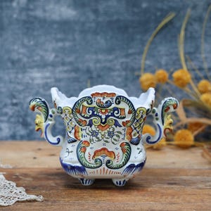 Puede incluir: Un jarrón de cerámica blanca con un diseño floral detallado en naranja, azul y verde. El jarrón tiene dos asas ornamentadas y se apoya en tres pequeños pies. El jarrón mide aproximadamente 15 cm de alto.