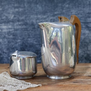 May include: A polished silver-toned metal pitcher with a light brown wooden handle and a matching lidded silver-toned metal sugar bowl. The pitcher features a curved spout and a hinged lid, while the sugar bowl has a small, rounded lid.