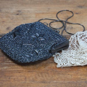 May include: A vintage, beaded evening bag in navy blue with a silver chain strap. The bag features a geometric pattern and is paired with a piece of white lace. The items rest on a wooden surface.