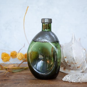 May include: A vintage green glass bottle with a metal cap. The bottle has an oval form and is placed on a wooden surface. A large seashell and dried yellow flowers are in the background.