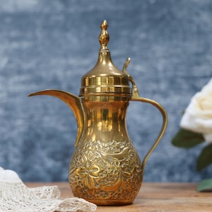 May include: A polished brass coffee pot featuring a long, slender spout, a curved handle, and a pointed lid. The lower body of the vessel is decorated with intricate, embossed floral patterns and textured detailing across the metallic surface.