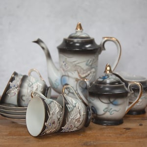 Puede incluir: Juego de té vintage con un esquema de color negro y dorado. El juego incluye una tetera, azucarero, lechera y varias tazas y platillos. La porcelana está decorada con un motivo de dragón y detalles dorados.