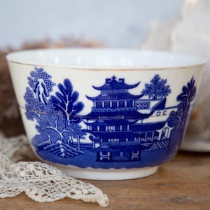 May include: A white ceramic bowl with a blue and white willow pattern. The bowl features a pagoda, trees, and a river.