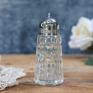 May include: A clear, vintage salt shaker with a silver-colored metal lid. The glass body has a textured, geometric pattern. The shaker is on a wooden surface, with a lace doily and white flowers in the background.