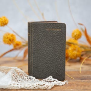 May include: A small, dark green New Testament book with gold lettering. The book has a textured cover and is resting on a wooden surface. A white lace cloth and dried yellow flowers are in the foreground. The background is blurred.