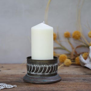 May include: A cream-coloured pillar candle in an antique brass candle holder. The holder features a detailed, patterned design. The candle and holder are on a wooden surface, with a seashell and dried flowers in the background.
