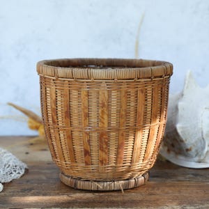 May include: A cylindrical woven basket, made from light brown and tan materials. The basket has a round base and is displayed on a wooden surface. The basket's height is approximately 20 cm.